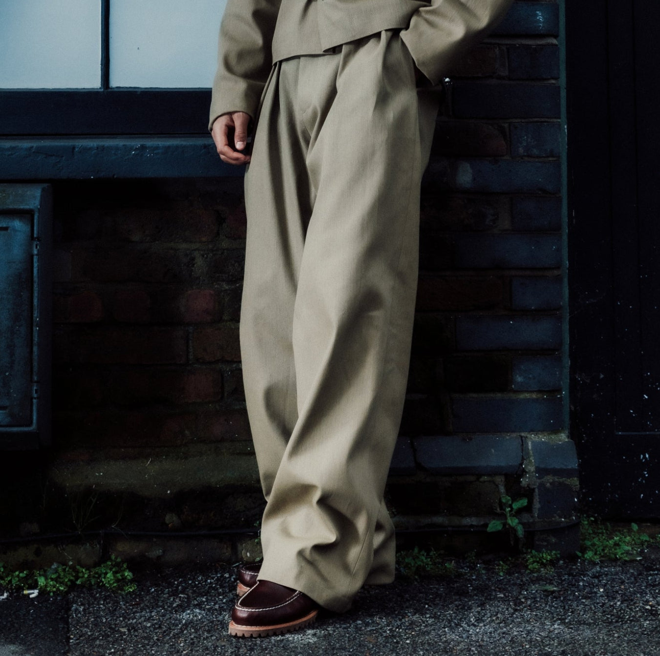 Person wearing a khaki outfit standing against a brick wall with a window in the background.