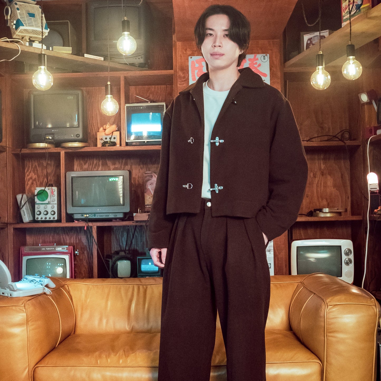 Person in a brown outfit standing in a room with vintage electronic equipment and a leather couch.