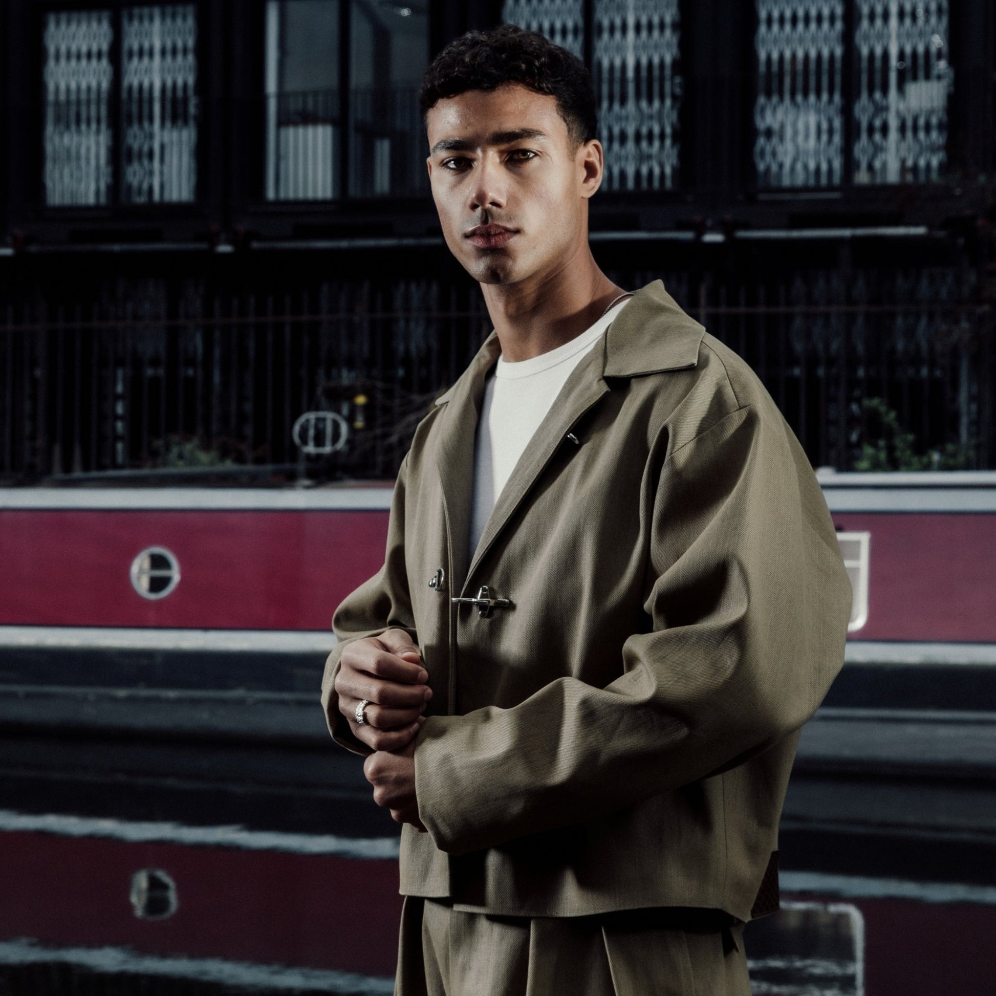 Man in a khaki cropped jacket standing in front of a building with a red train in the background