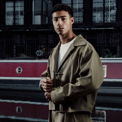 Man in a khaki cropped jacket standing in front of a building with a red train in the background