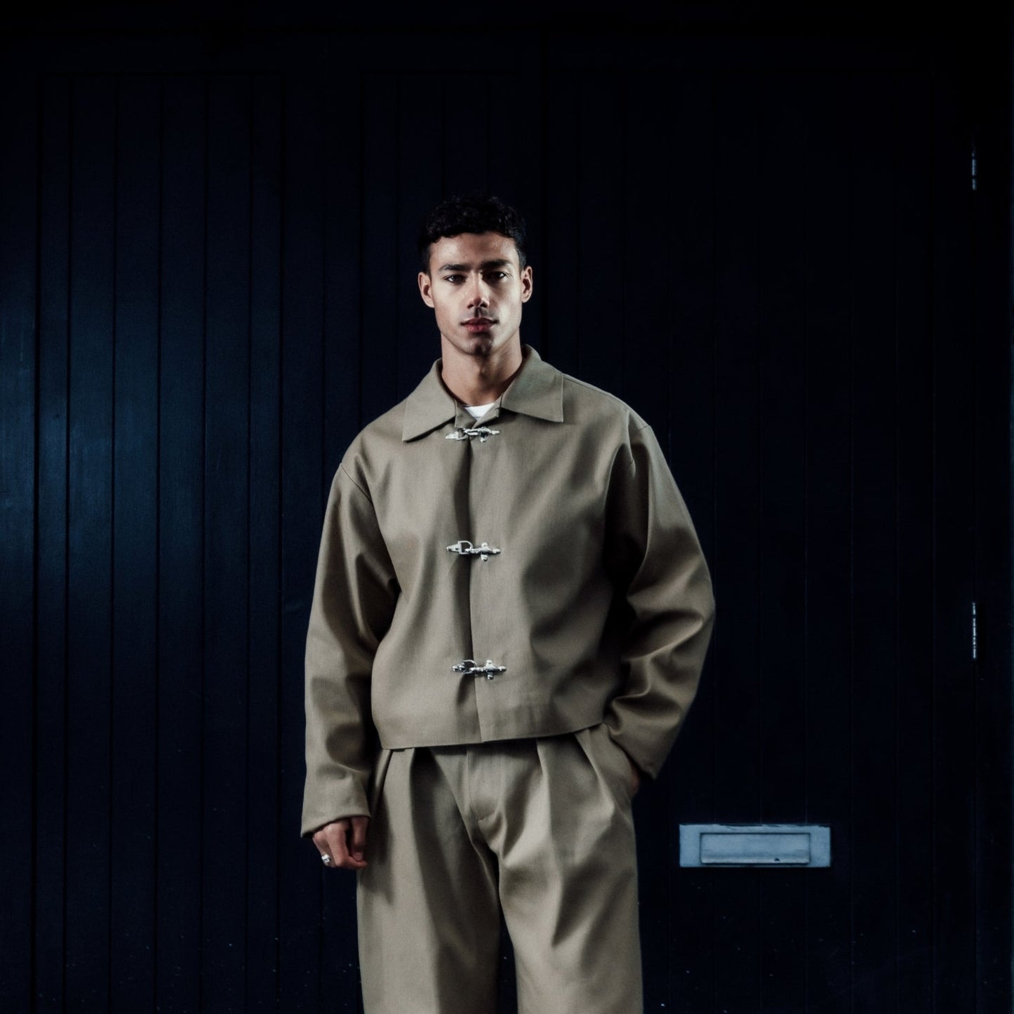 Man wearing a khaki outfit standing in front of a dark door on a brick wall.