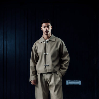 Man wearing a khaki outfit standing in front of a dark door on a brick wall.